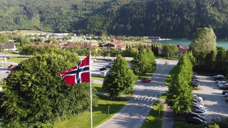 Norwegens-Flagge-Weht-Wunderschön-Im-Wind-Mit-Dem-Stadtzentrum-Von-Loen-Und-Dem-Fjord-Im-Hintergrund---Sich-Langsam-Bewegende-Luftaufnahmen-In-Wunderschöner-Landschaftsumgebung---Loen-Nordfjord-Norwegen
