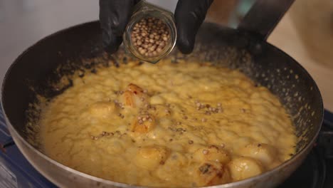 Professional-chef-adding-coriander-seeds-into-coconut-curry-scallops-seafood-dish,-cooking-on-the-pan,-simmering-to-brings-out-the-flavour-of-the-spices,-making-them-bloom-with-aromatic-smell