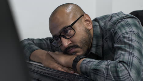 Close-up-shot-of-an-exhausted-young-Indian-entrepreneur-resting-his-head-on-his-arms-in-front-of-a-computer,-eyes-fixed-on-the-screen-waiting-for-confirmation-that-the-business-proposal-has-been-sent