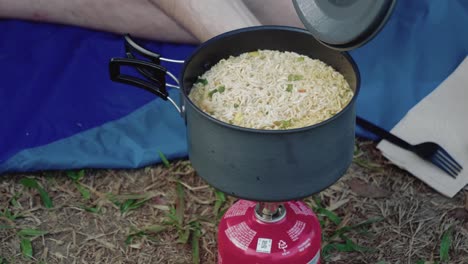 Man-opening-a-black-pot-filled-with-noodles-that-have-been-cooking-outdoors