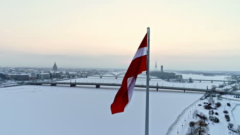 Toma-Aérea-En-órbita-De-La-Bandera-Letona-Y-El-Nevado-Río-Daugava-Durante-El-Frío-Día-De-Invierno-En-Riga