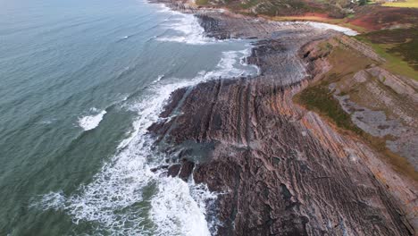 AERIAL:-Fly-towards-along-textured-rocky-coastline-with-crashing-waves,-Port-Eynon-Gower,-4k-Drone