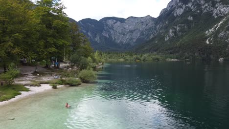 Lake-Bohinj-at-Gorenjska,-Slovenia---Aerial-Drone-View-of-Tourists-Relaxing-at-the-Waterfront-and-Swimming-in-the-Clear-Emerald-Lake
