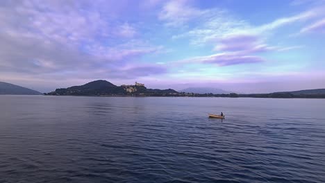 Wide-angle-view-of-small-fishing-boat-with-fisherman-rowing-in-calm-lake-waters-of-Maggiore-lake-in-Italy-with-Angera-castle-in-background