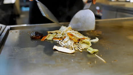 Un-Chef-Japonés-Que-Prepara-Verduras-Mezcladas-Salteadas-En-Una-Estufa-De-Placa-Caliente-Para-Cenar-En-Los-Clientes-En-Un-Restaurante-De-Ambiente