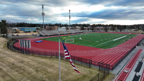 American-flag-waves-in-wind-at-athletic-football-soccer-field,-stadium-campus-aerial-view