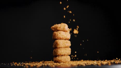 Stack-of-vegan-chicken-nuggets-with-bread-crumbs-falling-on-them-while-they-rotate,-slow-motion,-black-background,-static-shot