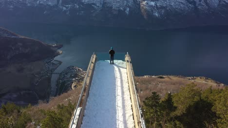 Aerial-along-walkway-of-Stegastein-viewpoint-with-man-standing-on-edge---Slow-tilt-down-to-reveal-hillside-forest-and-Aurland-village-beside-Aurlandsfjord---Sunny-day-Norway