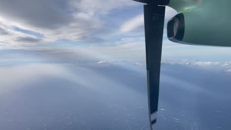Domestic-Aircraft-Norway,-turboprop-engine,-Filmed-out-of-window-above-arctic-northern-Norway,-with-snow-capped-mountings,-beams-of-light,-and-clouds,-Lofoten-Island-in-the-background,tilt-upward-shot