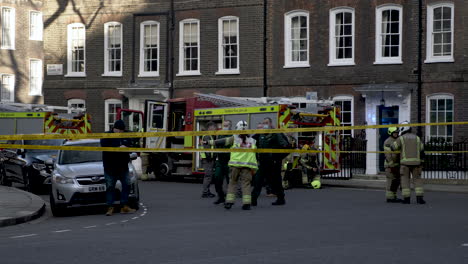 A-team-of-firefighters-at-the-scene-of-a-fire-in-Smiths-Square-London,-a-Fireman-leading-investigators-to-a-safe-location-on-the-perimeter-allowing-emergency-services-to-do-their-job,-England