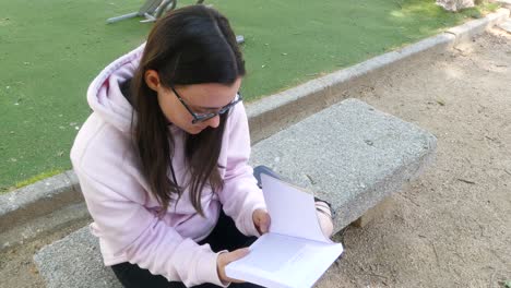 Young-and-cute-girl-with-glasses-reading-a-novel-on-a-stone-bench-in-a-park