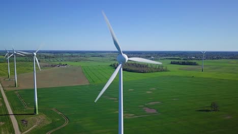 Aerial-view-of-wind-turbines-generating-renewable-energy-in-the-wind-farm,-sunny-spring-day,-low-flyover-over-green-agricultural-cereal-fields,-countryside-roads,-drone-dolly-shot-moving-left