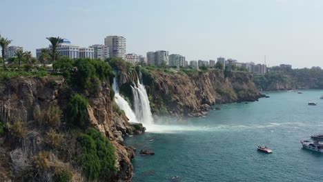 Luftdrohne-Passiert-Eine-Klippe-Und-Zeigt-Das-Wasser-Des-Unteren-Duden-Wasserfalls,-Das-Von-Einer-Felsigen-Klippe-Direkt-Ins-Mittelmeer-In-Antalya,-Türkei,-Fällt,-Während-Touristenboote-Unten-Blicken