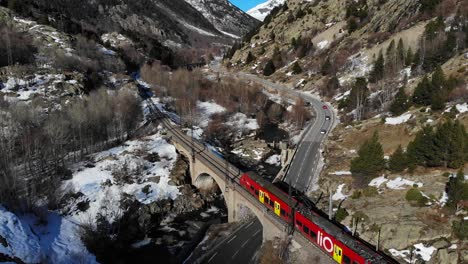 Luftaufnahme:-Roter-Zug-überquert-Eine-Straße-Mit-Wenig-Verkehr-Und-Einen-Fluss-Auf-Einer-Steinbrücke-In-Den-Pyrenäen-Zwischen-Schneebedeckten-Bergen