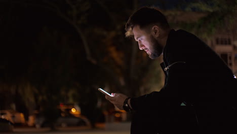 Night-time-medium-shot-of-a-man-sitting-on-a-bench-in-a-park,-scrolling-searching-on-a-smartphone