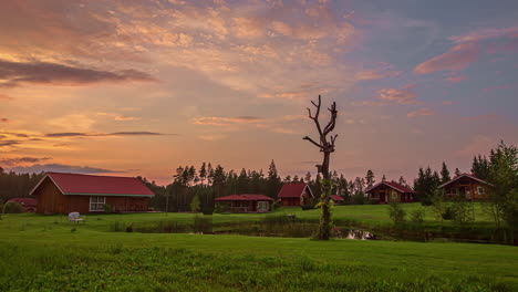 Red-Wooden-Cabins-With-Pond-And-Green-Grass-At-Sunset