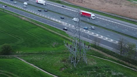 Vehicles-on-M62-motorway-passing-pylon-tower-on-countryside-farmland-fields-aerial-view-high-angle