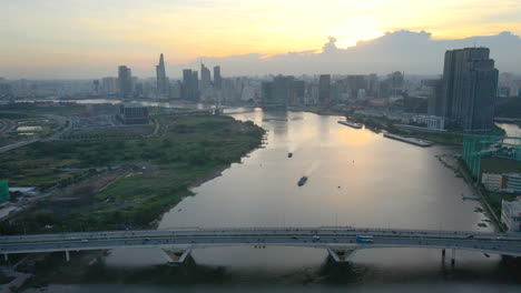 Flying-at-Sunset-over-Saigon-River-Bridge-near-Downtown-of-the-City-of-Ho-Chi-Minh-Vietnam,-Boats-Towers-and-Beautiful-Golden-Sky-in-Background