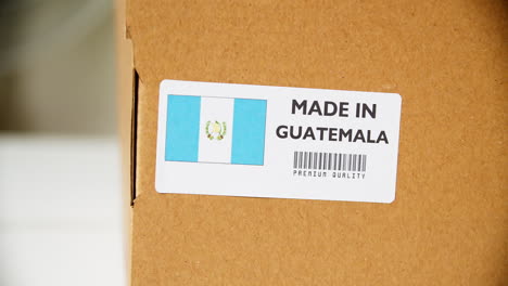 Manos-Aplicando-La-Etiqueta-De-La-Bandera-De-Guatemala-En-Una-Caja-De-Cartón-De-Envío-Con-Productos