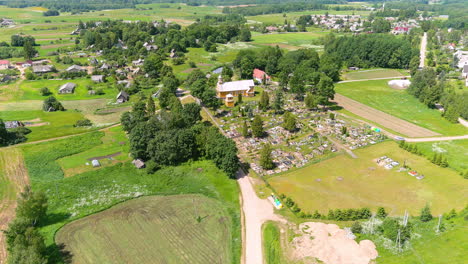 Aerial-View-of-Rural-Lithuanian-Village,-Catholic-Church,-Graveyard-and-Green-Landscape-on-Sunny-Summer-Day,-Drone-Shot