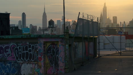 Muro-Con-Graffiti-Y-Cerca-De-Alambre-De-Púas-En-Queens,-Nueva-York,-Con-El-Horizonte-De-La-Ciudad-De-Nueva-York-Al-Atardecer-En-La-Distancia