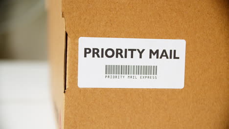 Hände-Kleben-Das-Aufkleberetikett-„Priority-Mail“-Auf-Einen-Briefkasten-Aus-Pappe-Mit-Barcode-Express