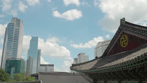 Fluffy-clouds-moving-over-the-roof-of-main-building-in-Bongeunsa-Temple-with-view-of-Trade-Tower,-Asem-Tower-skyscrapers-in-center-of-Seoul