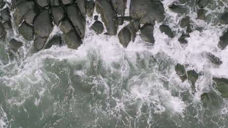 Slow-motion-aerial-overhead-top-down-footage-of-ocean-waves-crashing-into-rocks-that-are-on-the-coast