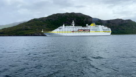 Low-medium-shot-of-the-cruise-ship-Hamburg-passing-through-the-arctic-circle-in-front-of-green-rocky-hills,-and-overcast-sky-in-Helgeland-Norway