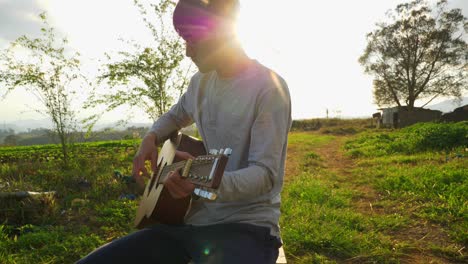 Asian-male-musician-playing-guitar-while-sitting-with-sun-shining-and-flares,-Vietnam