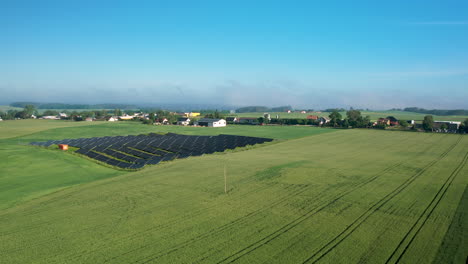 Sonnenkollektoren-Auf-Einem-Grünen-Feld-Mit-Dorfhäusern-Im-Hintergrund-Auf-Dem-Land-In-Polen---Schwenk-Aus-Der-Luft