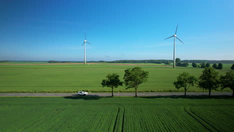 White-Car-Passing-on-Road-with-Wind-Turbine-in-Background,-Aerial-view