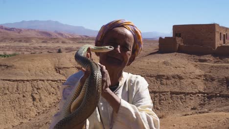 4K-Aufnahmen-Eines-Alten-Schlangenbeschwörers,-Der-Eine-Kobra-In-Der-Nähe-Der-Kasbah-Ait-Ben-Haddou-Im-Atlasgebirge-Von-Marokko-Hält