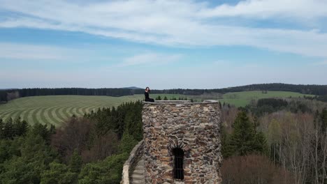 Aerial-View-of-Happy-Young-Woman-Sitting-on-Top-of-Medieval-Tower-and-Waving-to-Drone-Camera,-Revealing-Landscape-Shot-50fps