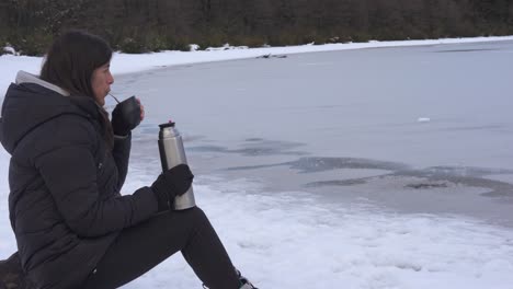 Eine-Frau-Trinkt-Etwas-Heißes-In-Einer-Sehr-Kalten-Umgebung-Mit-Schnee-Und-Zugefrorenem-See