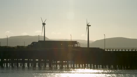 Wunderschöne-Aufnahme-Von-Windkraftanlagen,-Die-Sich-Bei-Sonnenaufgang-Drehen,-Und-Erstaunlichen-Sonnenstrahlen-Und-Spiegelungen-Auf-Dem-Wasser-In-Swansea-Marina,-Großbritannien,-4K