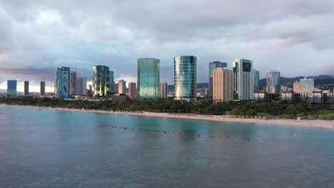 Aerial-wide-rising-shot-of-the-sun-reflecting-off-buildings-in-downtown-Honolulu,-Hawaii-at-sunset