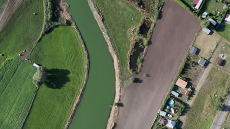 Aerial-top-down-shot-of-a-curvy-river-running-among-the-fields,-farms-and-gardens-of-a-countryside-farming-village
