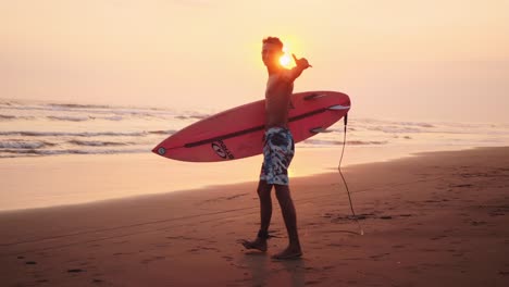 Der-Gutaussehende-Surfer-Trägt-Das-Brett-Zum-Wasser,-Um-Das-Shaka-Brah-Sonnenaufgangssignal-Zu-Signalisieren
