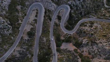 Aerial-drone-top-down-shot-over-cars-passing-along-winding-Sa-Calobra-road-over-rocky-mountain-range-in-Mallorca,-Spain-during-evening-time