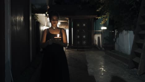 Front-view-of-woman-in-black-dress-walking-down-dark-hallway-holding-a-burning-candle-in-hands