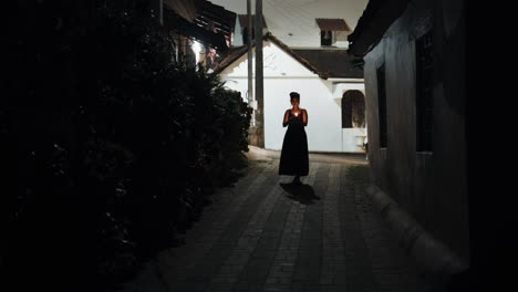 Front-view-of-pretty-woman-in-black-dress-walking-along-dark-street-over-mosaic-floor-holding-lit-candle-in-hands-while-dog-looking-at-her-in-background