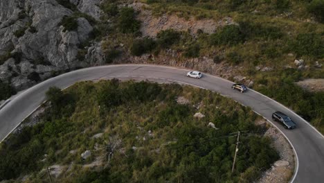 Aerial-drone-shot-over-cars-passing-by-at-a-U-turn-of-winding-Sa-Calobra-road-over-rocky-mountain-range-in-Mallorca,-Spain-during-evening-time