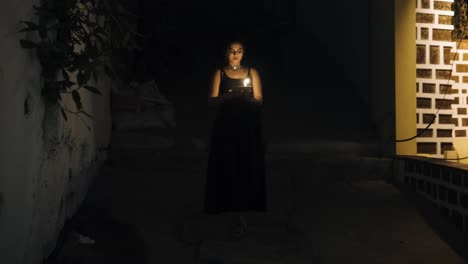 Front-view-of-woman-in-black-dress-walks-down-the-dark-stairs-holding-a-burning-candle-in-her-hands-with-bushes-nearby