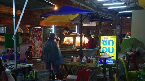 Vendedor-De-Comida-Con-Clientes-En-Una-Calle-Concurrida-En-Tailandia-Durante-La-Noche-Lluviosa-Y-Tormentosa