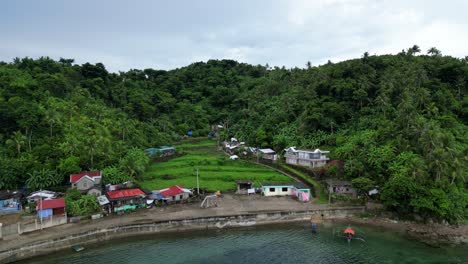 Aerial-Rotating-View-of-Idyllic,-Coastal-Tropical-Village-with-lush-rainforest-covered-hill-in-background