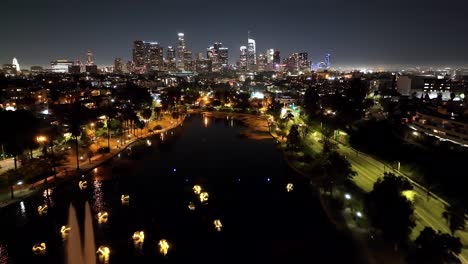 Aerial-view-flying-over-Los-Angeles-Echo-park-illuminated-lake-fountain-towards-futuristic-city-skyline