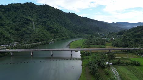Stunning-Aerial-View-of-Busy-Bridge-crossing-vast,-quaint-river-with-lush-rainforests-and-hills-in-background