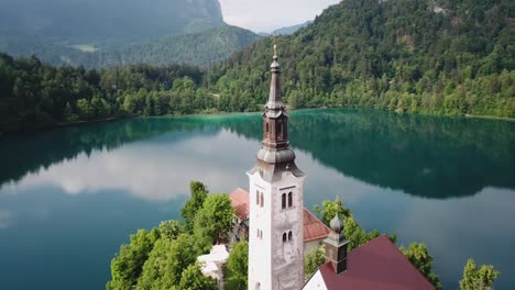 Orbiting-aerial-view-of-a-church-tower-surrounded-by-Lake-Bled's-clear-water-in-Slovenia