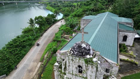 Cinematic,-Establishing-Aerial-View-of-local,-riverside-Catholic-Church-alongside-tricycles-driving-on-road-and-lush-rainforests